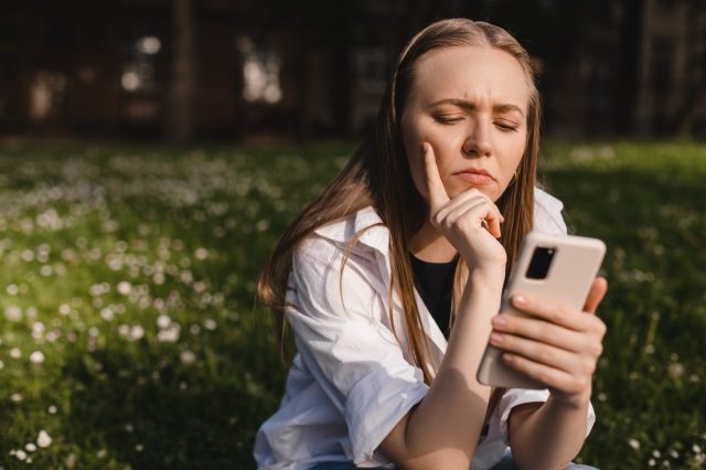 Woman holding a phone while holding her hand on chin. Expressive student thinking outside in campus - Woman in doubt with smartphone in her palm, think about reading information, search solution.