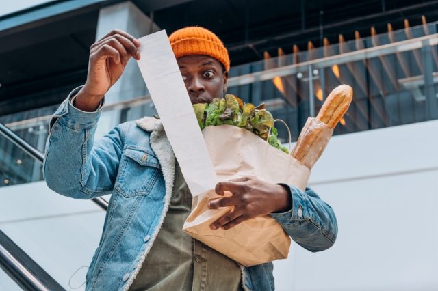 Doubting African-American person in denim jacket looks at sales paper receipt total holding pack with food products on escalator