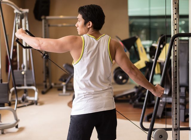 Rear view of a strong young man lifting cable with single arm, while exercising lateral or side raise for shoulder muscles in a modern fitness club