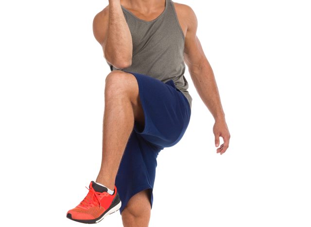 Smiling young fit man in gray tank top, blue shorts and orange sneakers is standing on one leg and touching with his elbow to the knee. Full length studio shot isolated on white. Standing knee to elbow. 