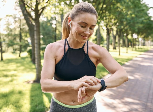 Morning jog. Beautiful sporty woman in sportswear and earphones looking at her watch, checking the result while running in a green park on a sunny summer day