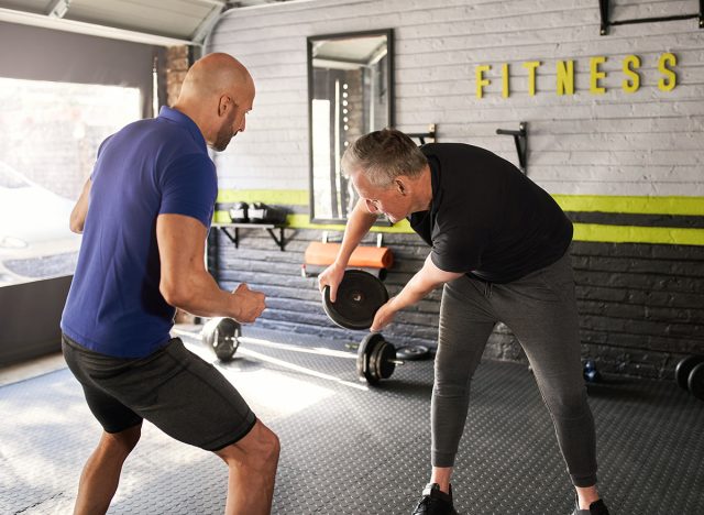 Male personal trainer motivating and supporting elderly senior man while doing wood choppers dynamic movement exercise in home gym small business