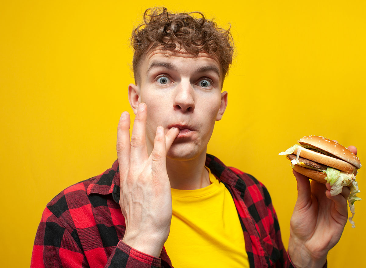 young surprised guy student eats a big tasty burger on a yellow background and licks the sauce on his finger, a man eats fast food and looks at the camera