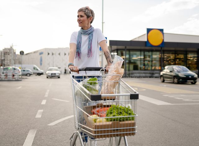 One mature woman caucasian female walking in the parking lot in front of the shopping center grocery store supermarket pushing chart happy smile half length copy space