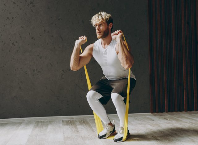 Full body side view young strong sporty athletic sportsman man wears white tank shirt black shorts using elastic rubber band for hands do squat warm up training indoor at gym. Workout sport concept. Resistance band squat. 