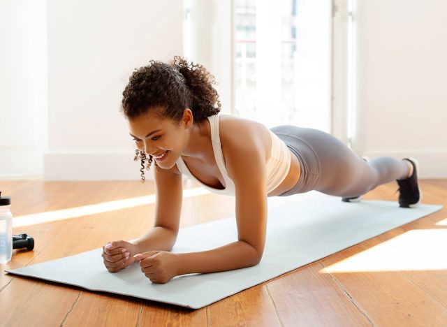 Young fitness woman in sportswear doing elbow plank exercise for core strengthening on fitness mat in modern living room at home interior. Healthy domestic workout concept, plank hold. 