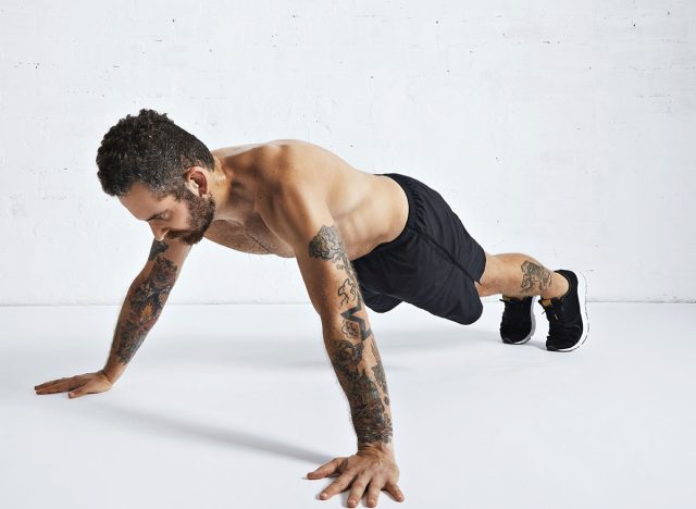 Strong tattoed and ripped male athlete shows how to do wide or archer push-ups, isolated on white spacious room, top position