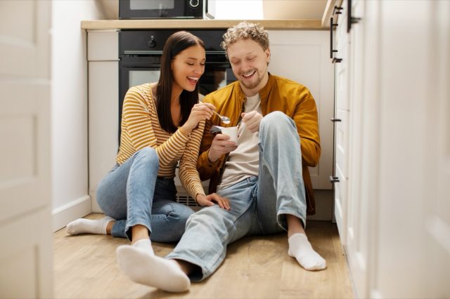 Happy couple sitting on kitchen floor, enjoying time together at home, eating yogurt or dessert. Marriage with romance