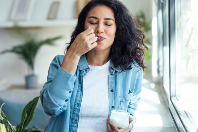 Shot of happy beautiful woman eating yogurt while standing in living room at home.