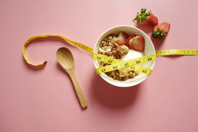 Healthy eating plan to lose weight theme yogurt bowl with granola and fresh fruits toppings wrapped around with yellow measuring tape on pink background with strawberries and wooden spoon. (top view)